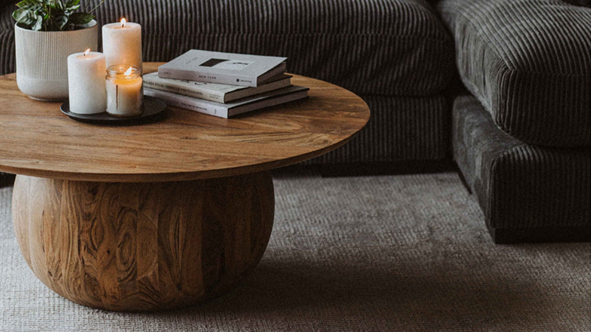 Moe's Home Collection Bradbury round coffee table in Natural acacia, the grain of its carved sphere base visible beside a dark corduroy sectional, with candles and books on the top