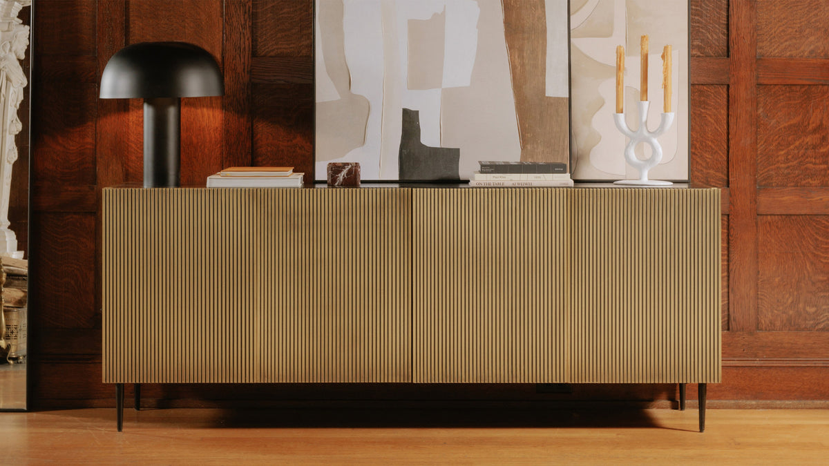 Moe's Home Collection Brogan sideboard in antique brass, its fluted doors and black top set against a dark panelled wall, with a domed black table lamp, candlesticks and small sculptures arranged across the top