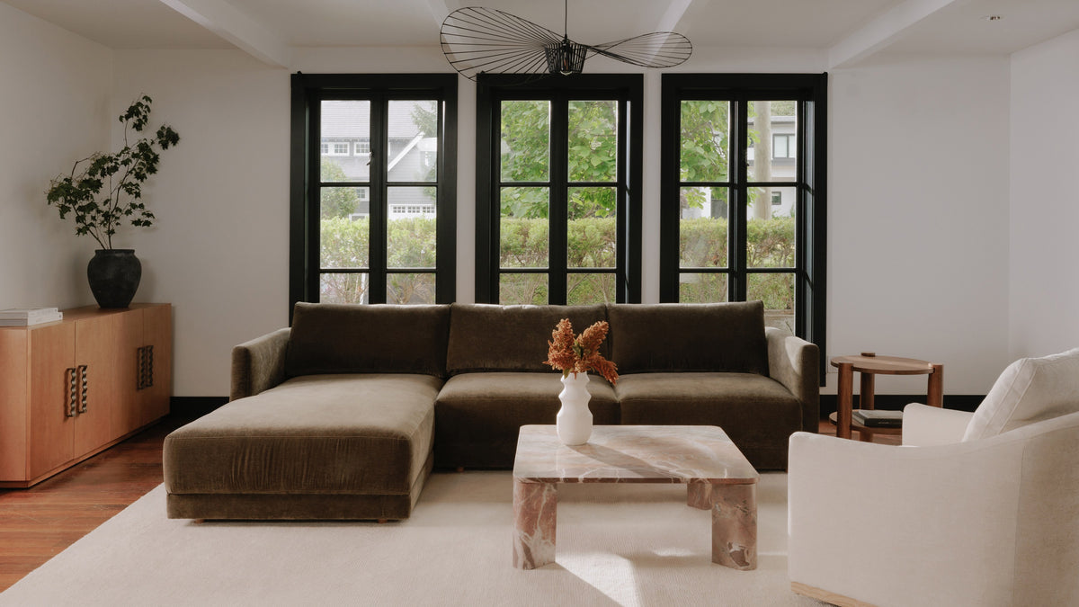 Moe's Home Collection Bryn sectional in heritage cedar green velvet with a left-facing chaise, seen straight on in a pale room with three black-framed windows behind it, a tan leather sideboard to the left and a marble coffee table in front