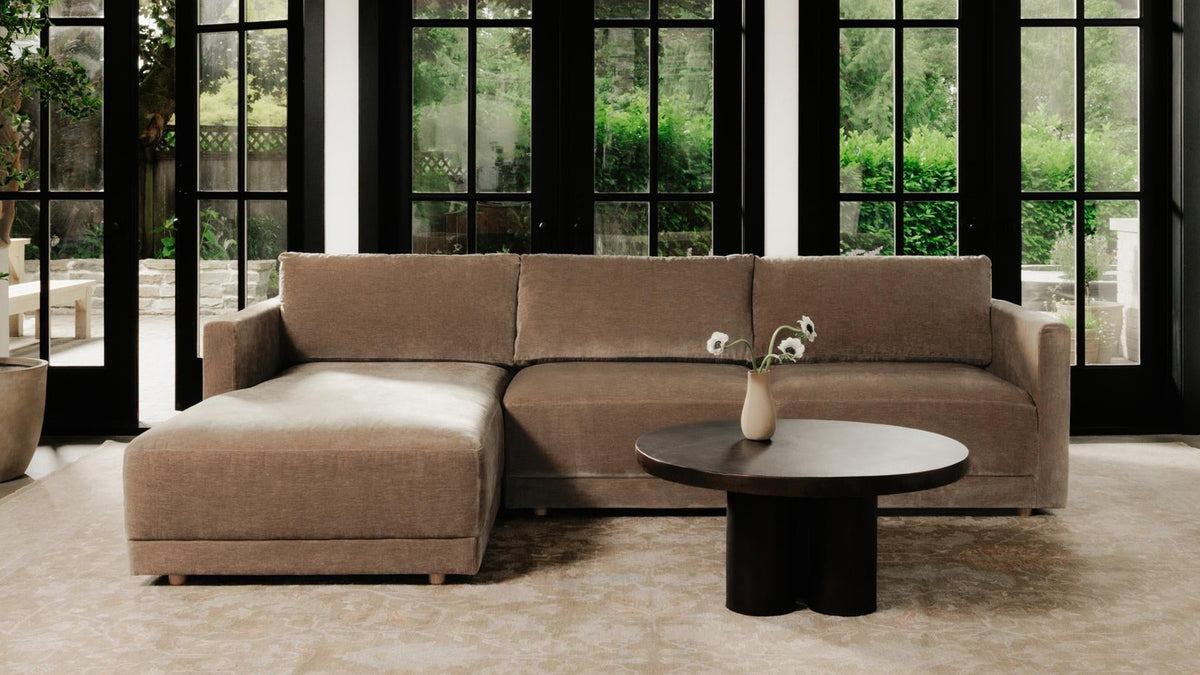 Moe's Home Collection Bryn sectional in camel chenille with a left-facing chaise, set on a faded rug in a garden room walled with black steel windows, an olive tree in a stone pot to the left and a round black coffee table in front