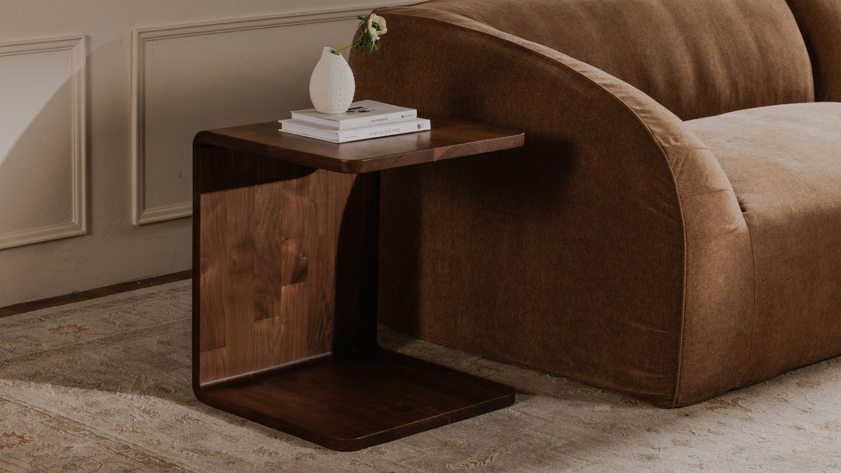 Moe's Home Collection Carrie side table in solid walnut, its C-shaped base slid under a caramel sofa with books and a small white vase on the top