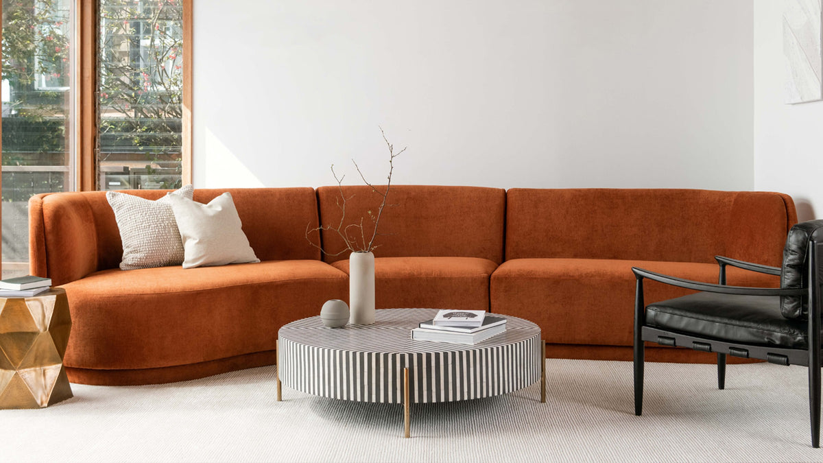 Moe's Home Collection Chameau round coffee table in black and white bone veneer, its striped drum lifted on slim brass legs before a rust curved sofa in a sunlit white room