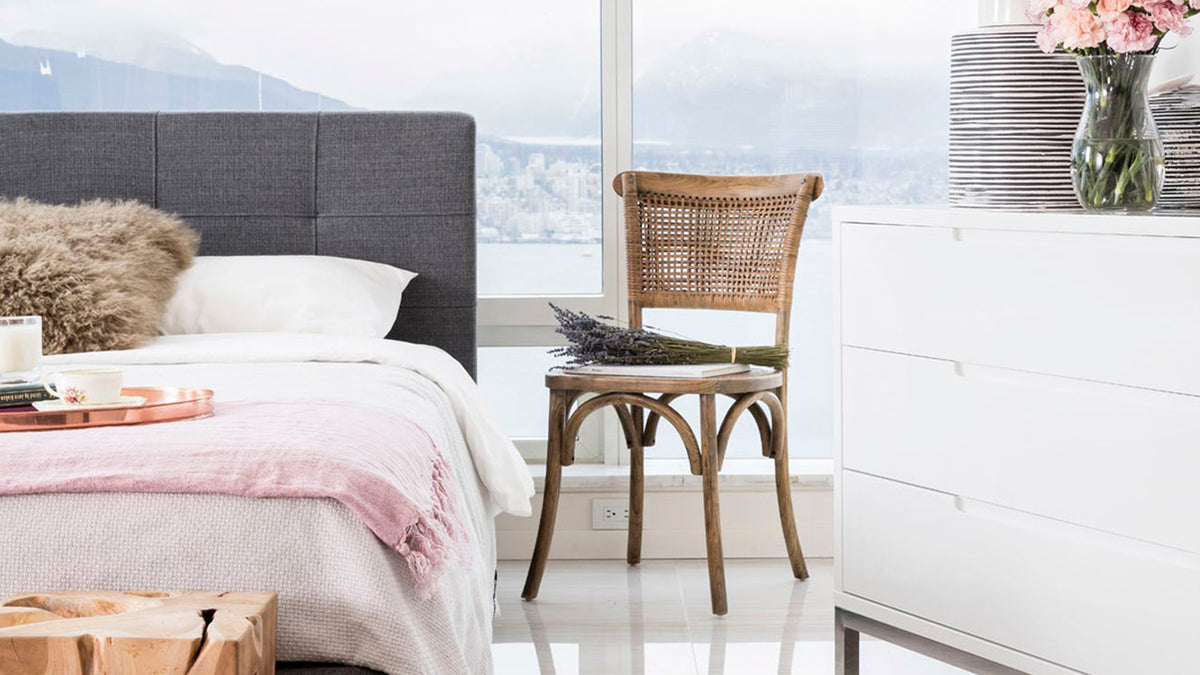 A Moe's Home Collection Churchill chair in light brown elm with a woven rattan back, beside a bed as a bedside seat, in front of a window looking over water and mountains