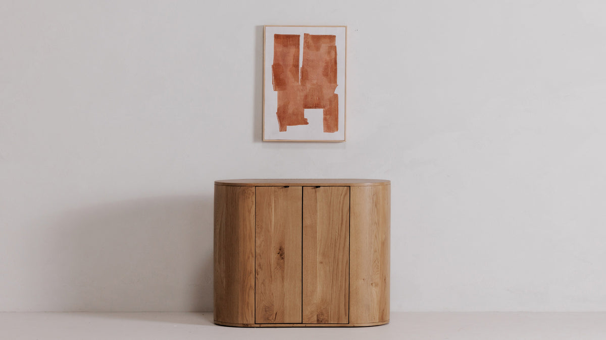 The Cici framed painting centered on a plain wall above a rounded oak cabinet, its rust-toned abstract shapes on white canvas in a pale pine frame