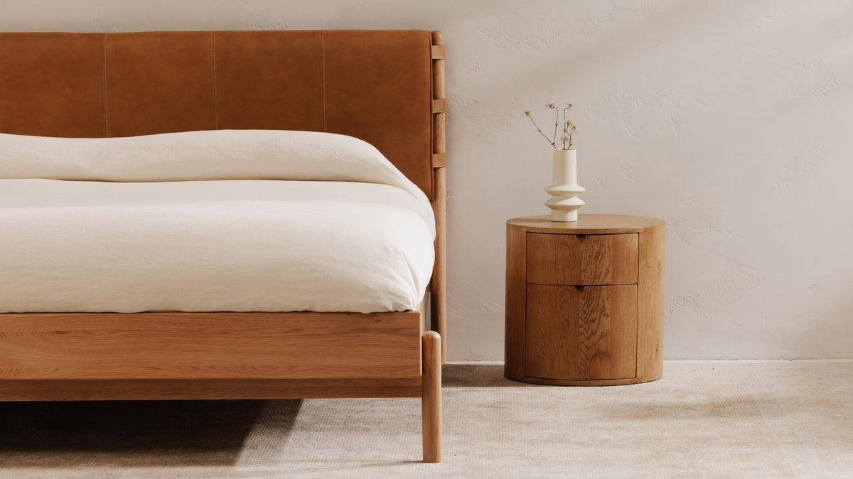 Close view of the Colby bed's leather headboard, oak side rail and rounded oak leg, with a round oak nightstand holding a white vase of dried flowers