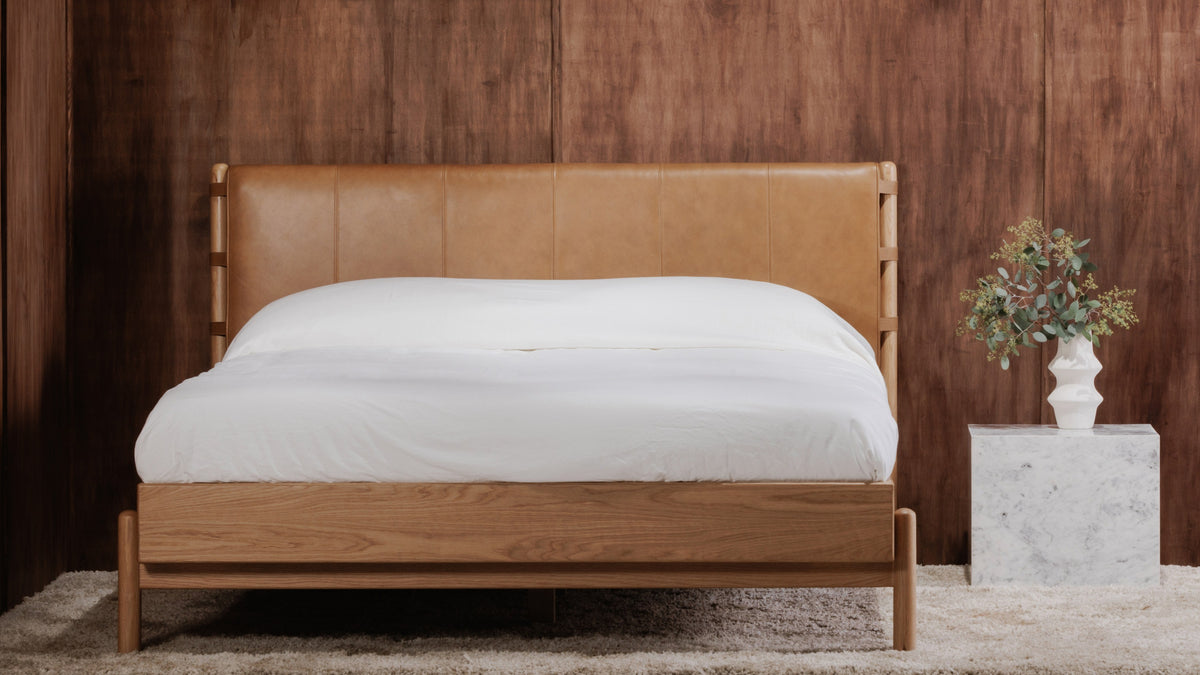 Colby bed in tan leather and oak on a cream shag rug in front of a walnut-panelled wall, with a marble cube side table and a vase of eucalyptus