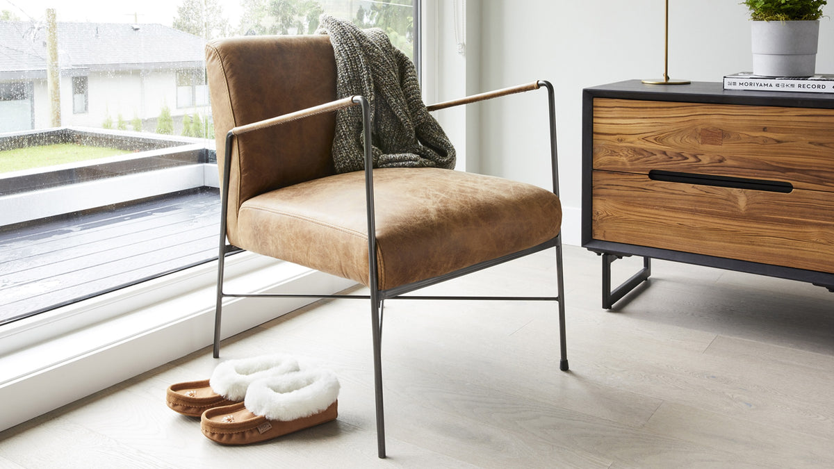 A Moe's Home Collection Dagwood armchair in brown top-grain leather on a slim dark iron frame, standing on pale wood flooring beside a full-height window, with a chunky gray knit throw over one shoulder of the back, sheepskin-lined slippers on the floor and a walnut and black media console to the right