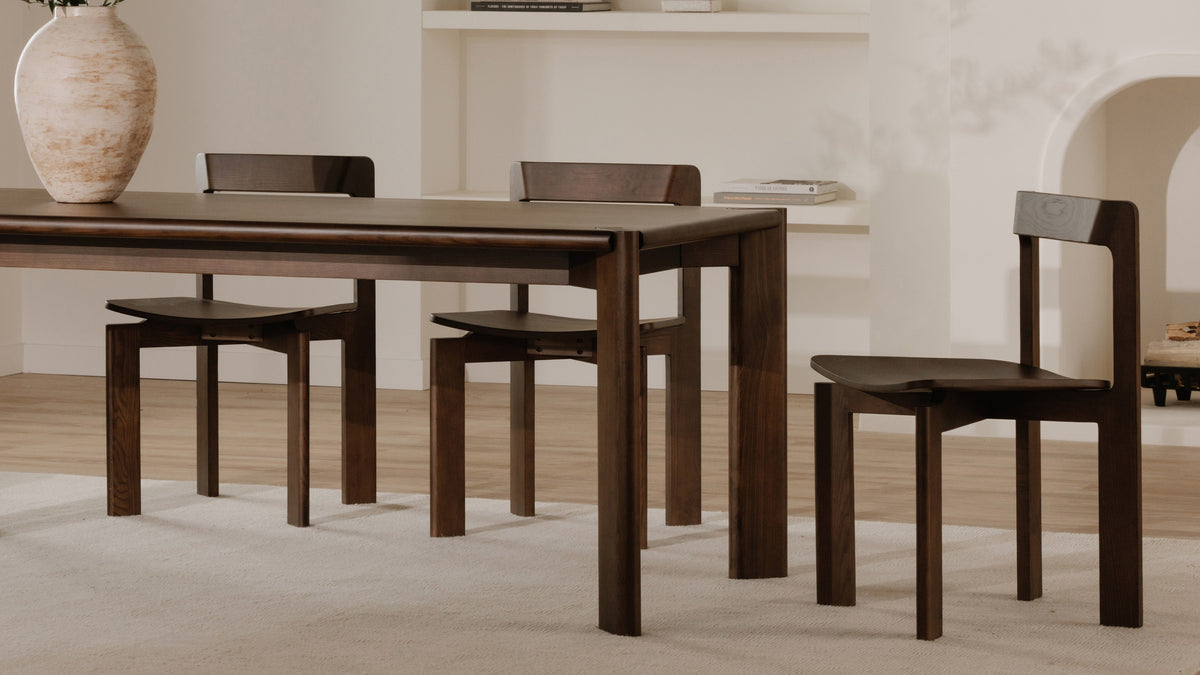 A close view of Moe's Home Collection Daifuku dining chairs in walnut-stained ash pulled up to a dining table, one chair in profile showing the shaped seat and low back