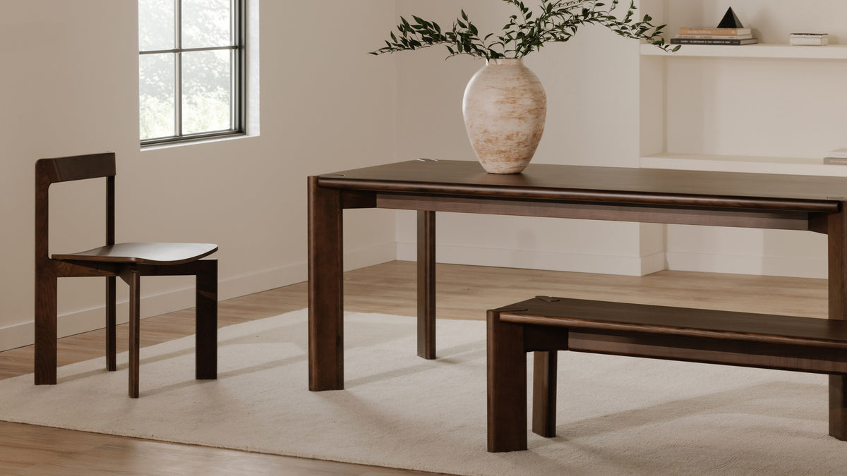 Daifuku dining table in walnut stained ash on a cream rug, with a matching bench and a single chair, a travertine vase of olive branches on the top and a window to the left