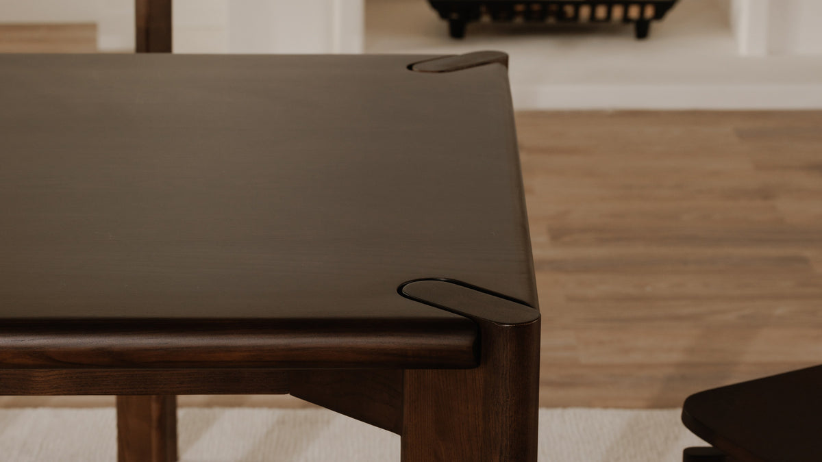 Close view of the Daifuku dining table corner in walnut stained ash, showing the rounded edge band interrupted at the corner and the top of the leg finishing flush with the tabletop