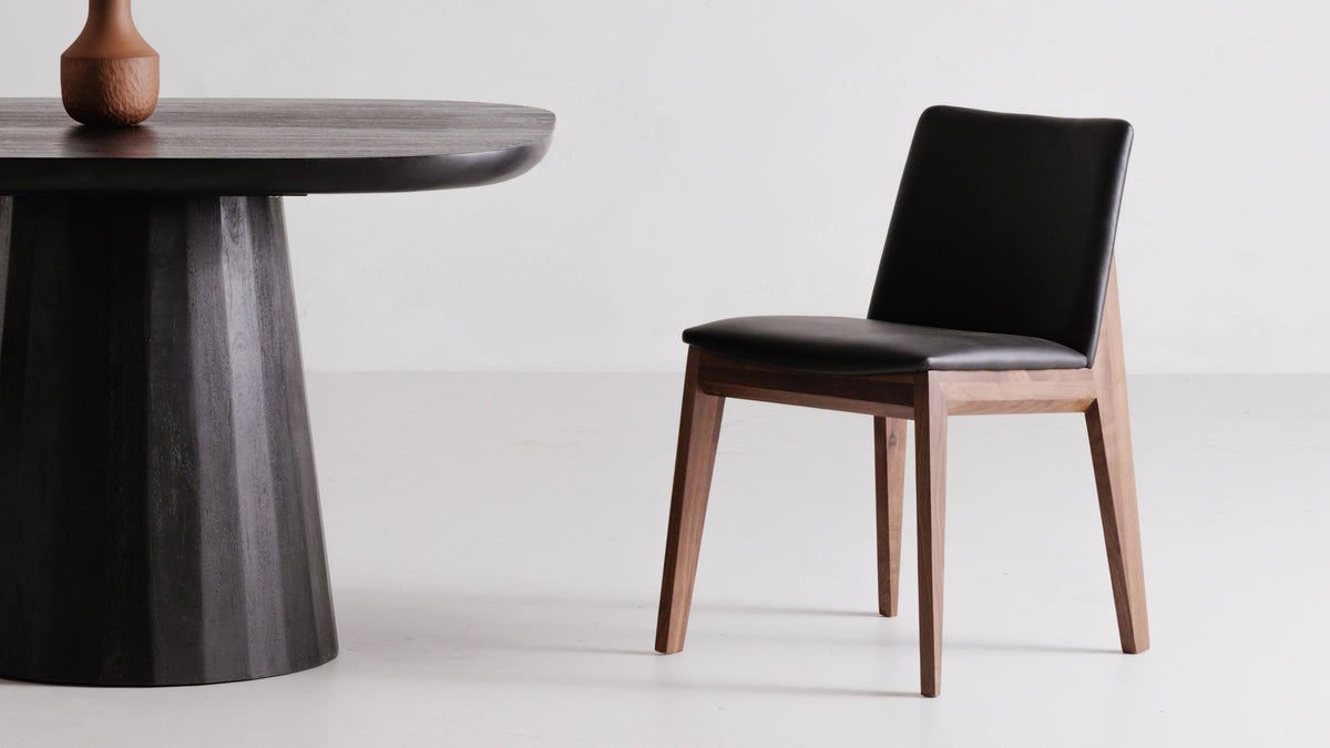 A single Moe's Home Collection Deco dining chair in ebony vegan leather beside a black round pedestal table in a white studio, showing the tapered walnut legs and angled seat