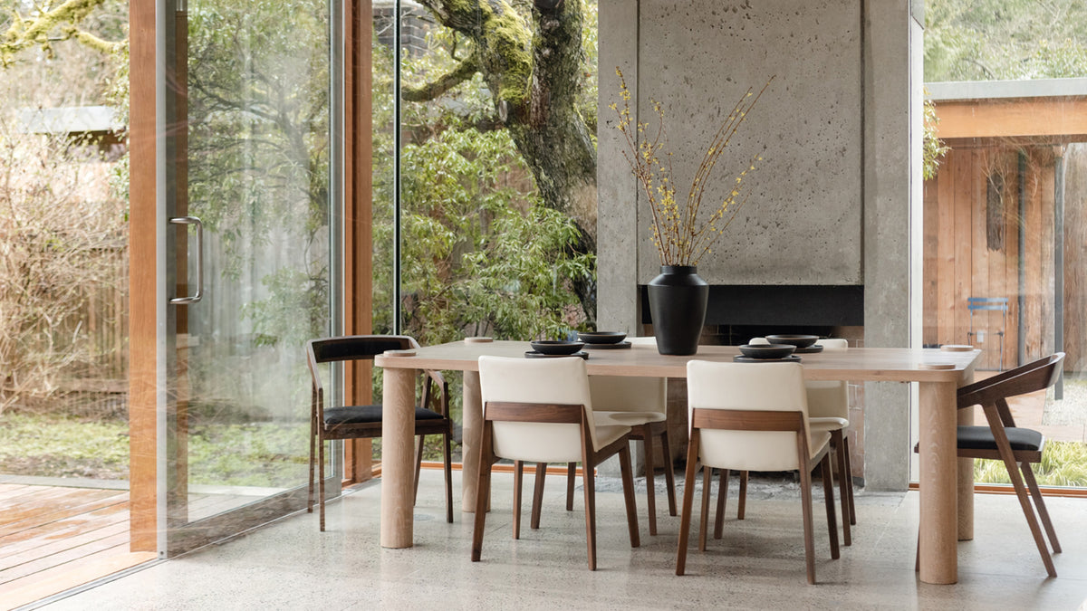 Moe's Home Collection Deco dining chairs in cream white vegan leather and black around a pale oak dining table in a glass-walled room, with a board-formed concrete fireplace and a garden beyond