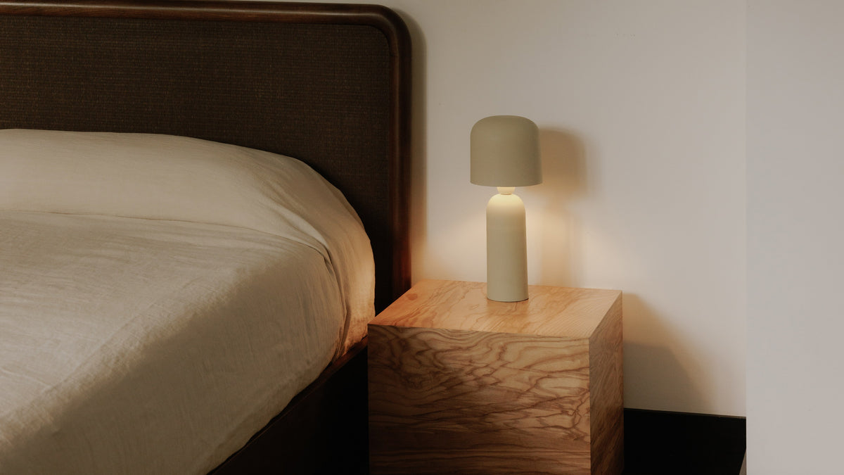 The Echo table lamp in cream, lit, its softly domed iron shade glowing on a burl-wood cube nightstand beside a bed with a caned headboard and linen bedding