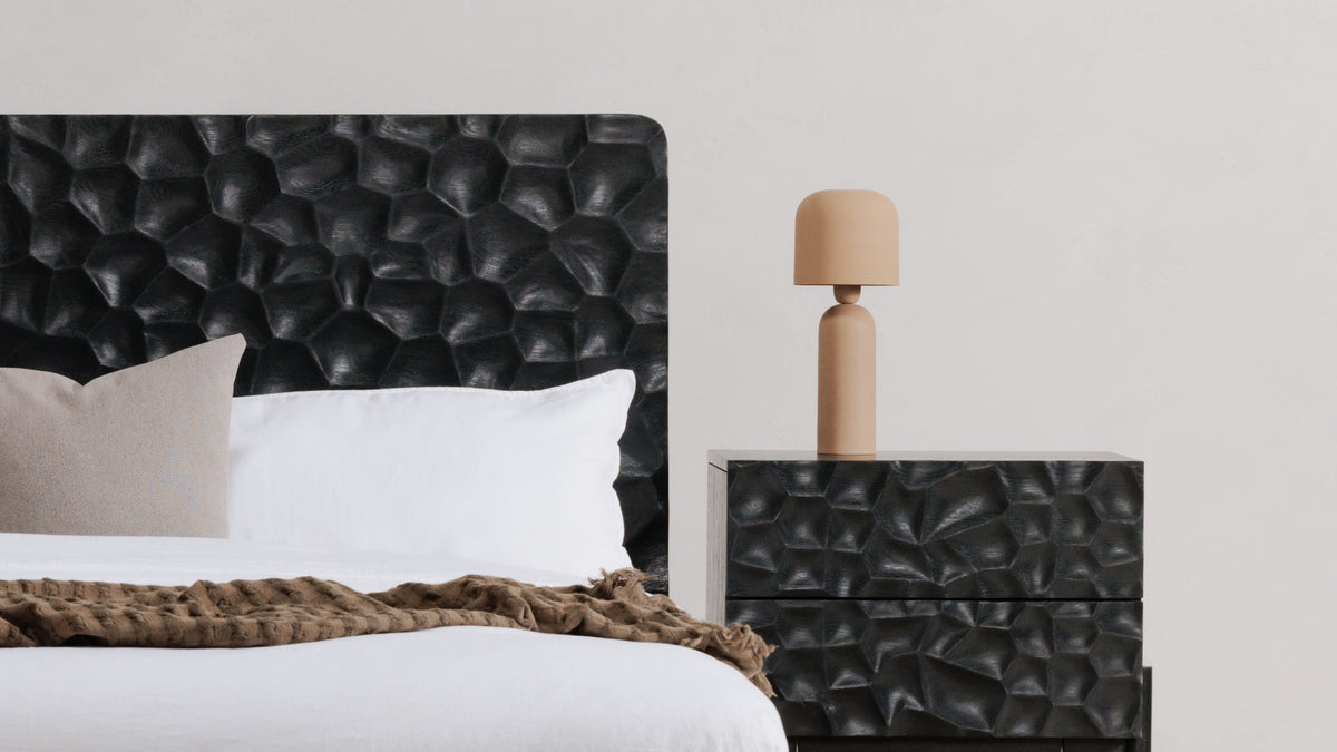 The Echo table lamp in terracotta on a faceted black nightstand beside a black leather headboard, white bedding and a brown knit throw