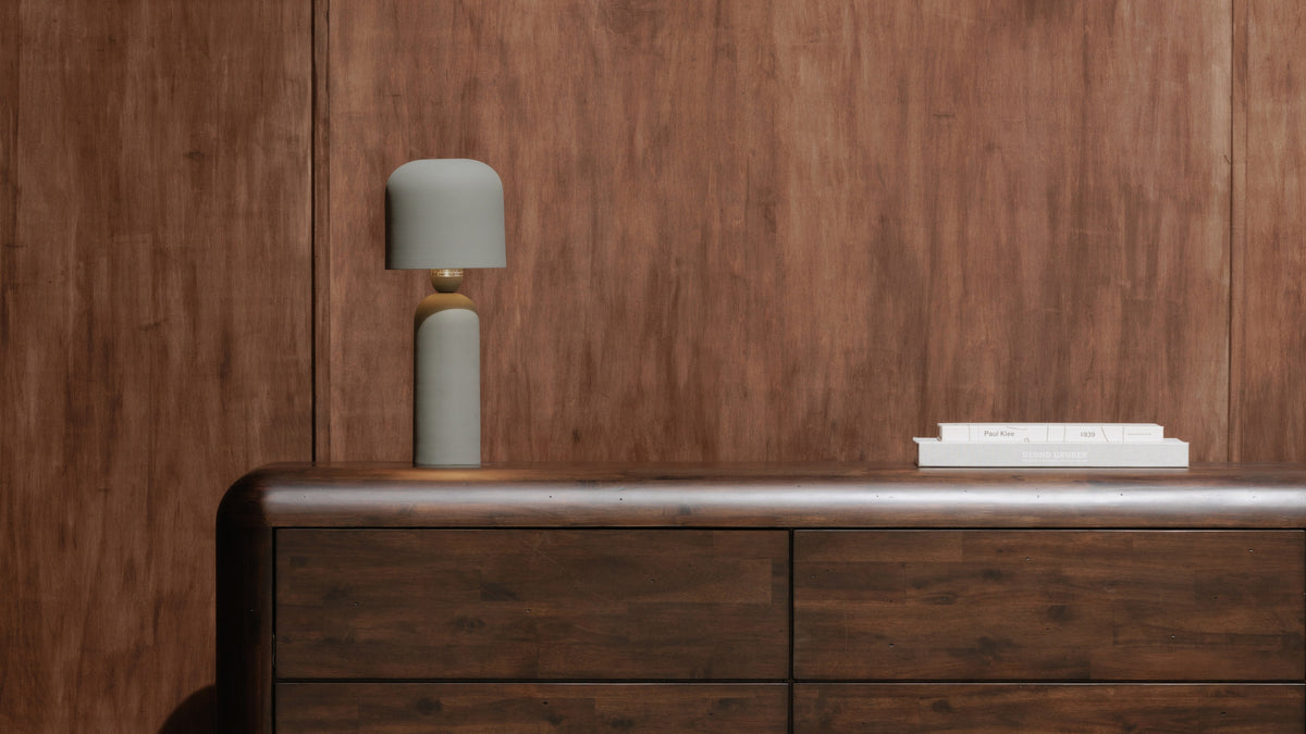 The Echo table lamp in green, lit, on a walnut dresser against a wood-panelled wall with a stack of white art books
