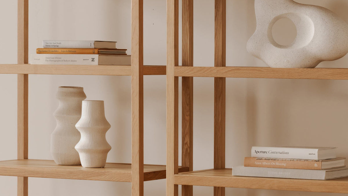 Two Moe's Home Collection Eero bookcases in natural oak seen close, the adjacent uprights meeting at the centre, with books and pale ceramics on the oak shelves