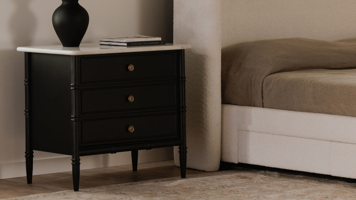 Moe's Home Collection Eleanor nightstand in matte black beside a boucle bed, its white Banswara marble top above three drawers with antique brass ring pulls, standing on slender turned legs with a black vase and books on top