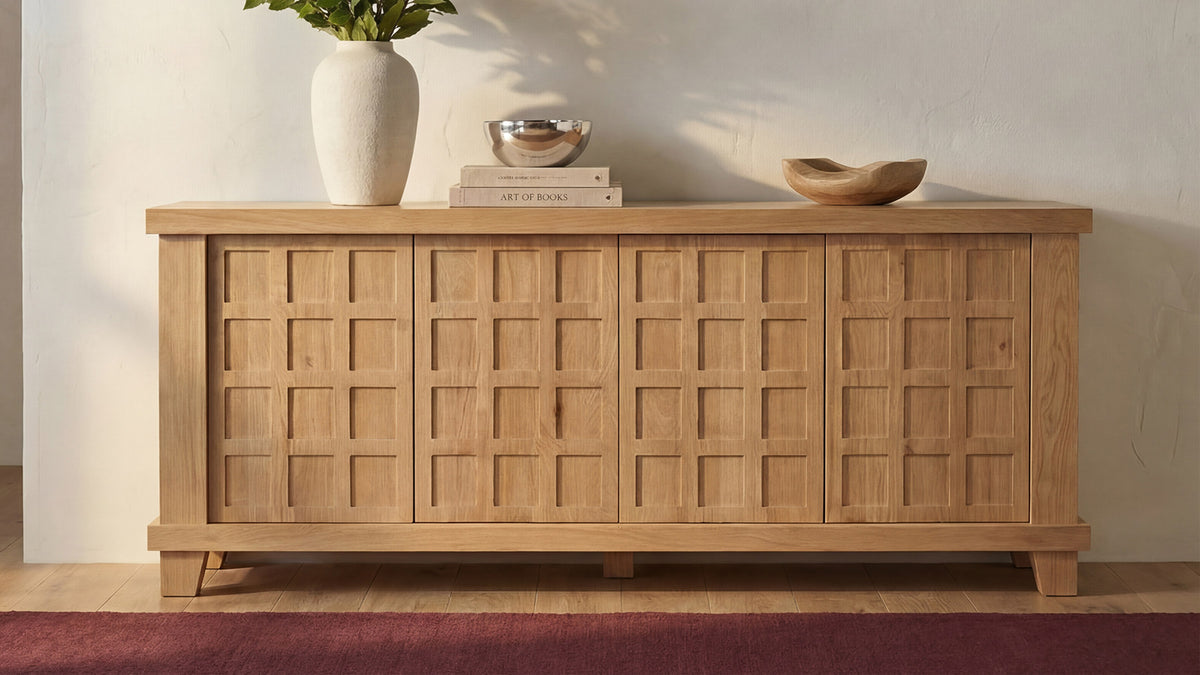 Moe's Home Collection Enya sideboard in oak against a plaster wall, its carved grid door fronts in raking sunlight, with a white vase, a stack of books and a shallow wooden bowl arranged on top