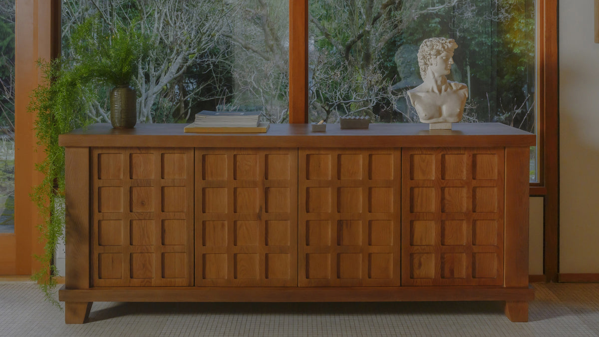 Moe's Home Collection Enya sideboard in oak beneath a window looking onto greenery, its carved grid doors seen straight on, with a classical bust and a planter on the sill above