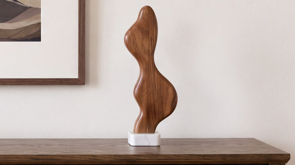 The Eon sculpture on a dark wood surface, its curving acacia form on a white marble base, with a framed landscape resting against the wall behind