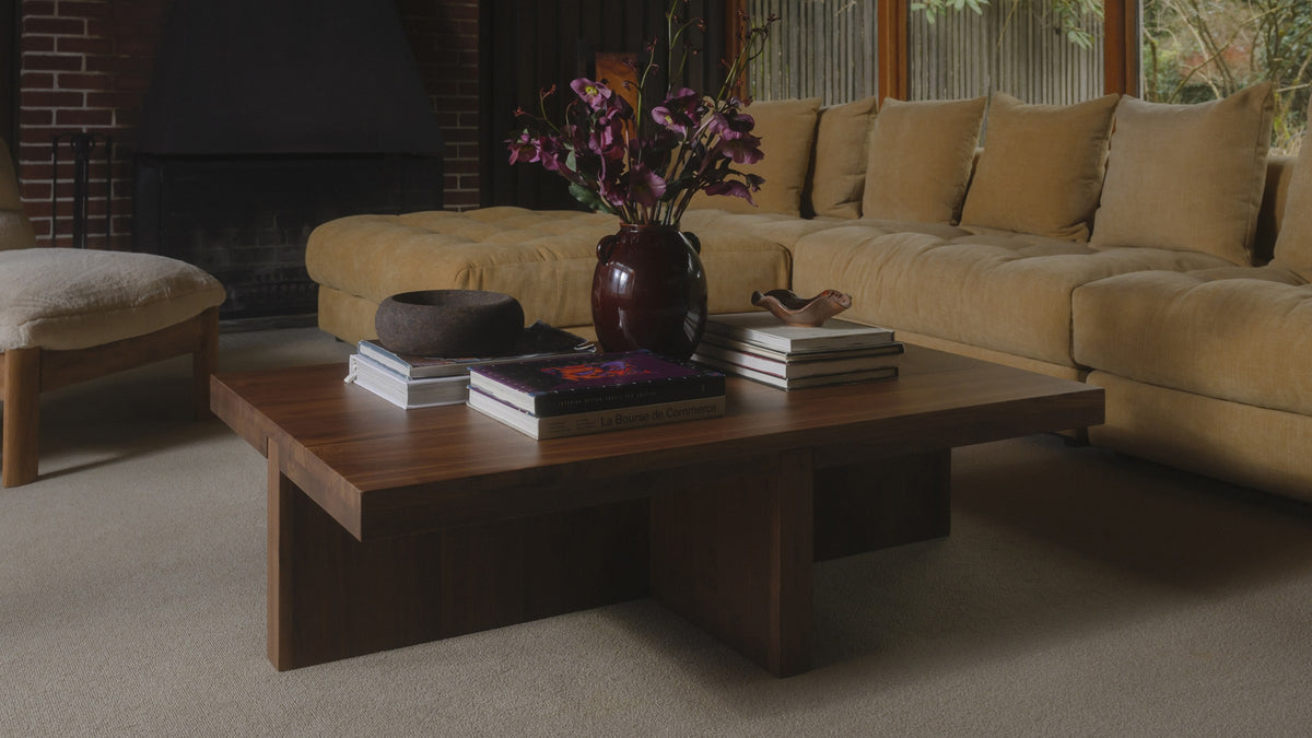 Moe's Home Collection Folke rectangular coffee table in solid walnut seen from the side, its T-shaped base clearly visible, in a panelled room with a brick fireplace