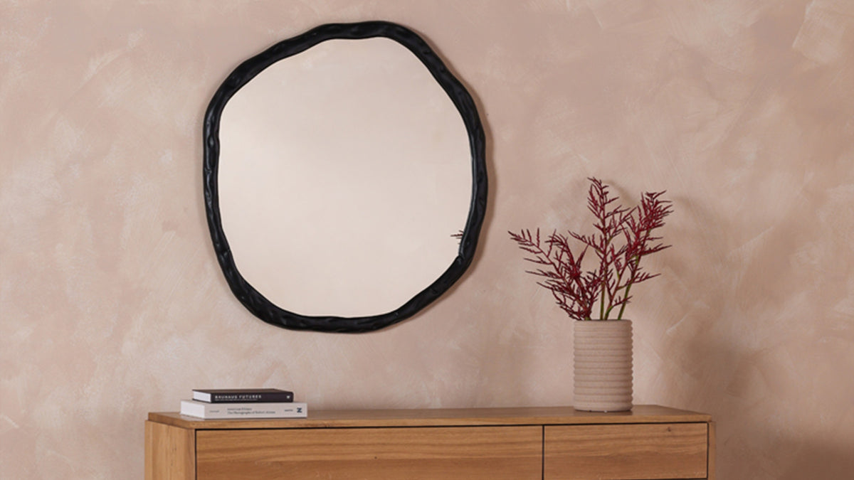 The large Foundry mirror in black above an oak console, its irregular cast edge catching the light, beside a ribbed vase of dried red stems