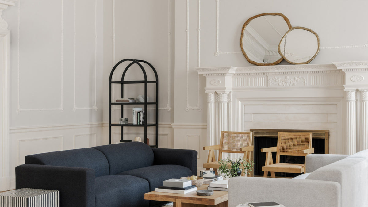 Two gold Foundry mirrors overlapping above a white fireplace in a paneled room, with a navy sofa and a black shelving unit in the foreground