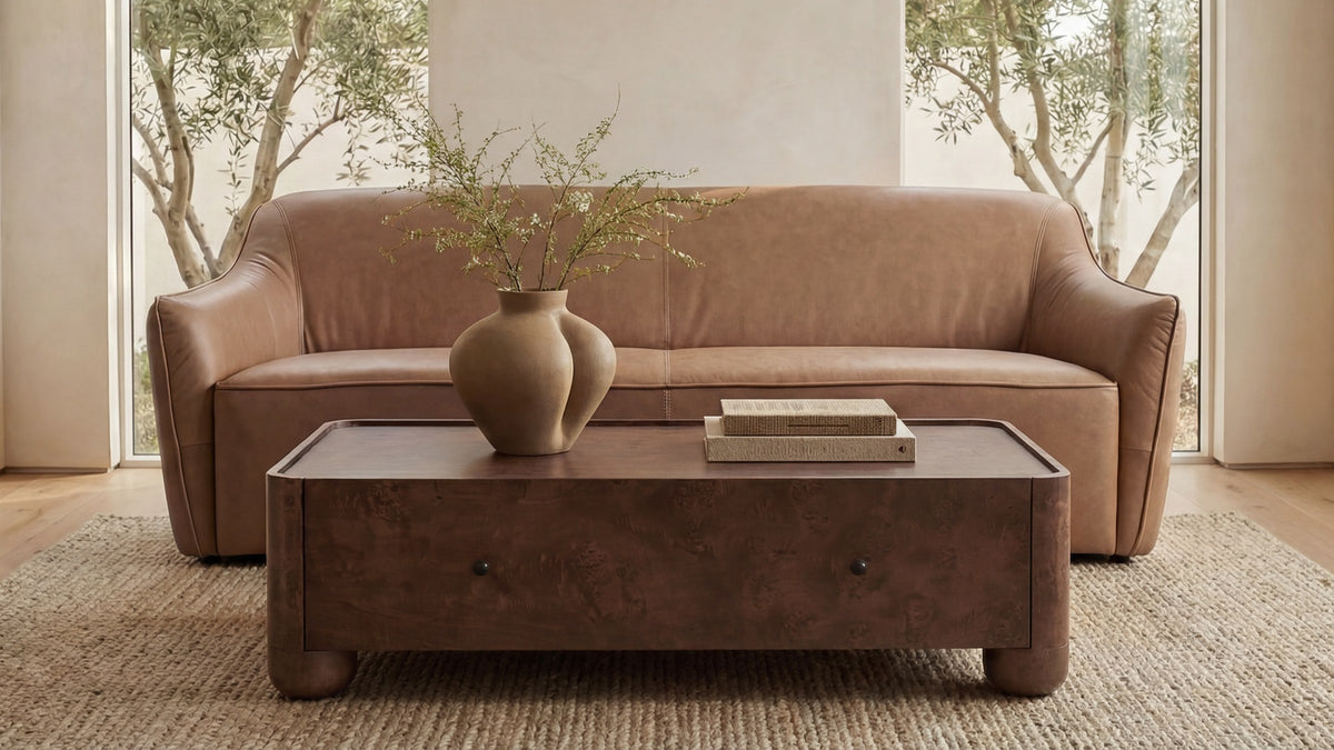 Moe's Home Collection Gibson coffee table in the Burl finish, its figured dark veneer and black metal knobs set against a dusty-rose leather sofa on a jute rug, with a ceramic vase and two books on the top