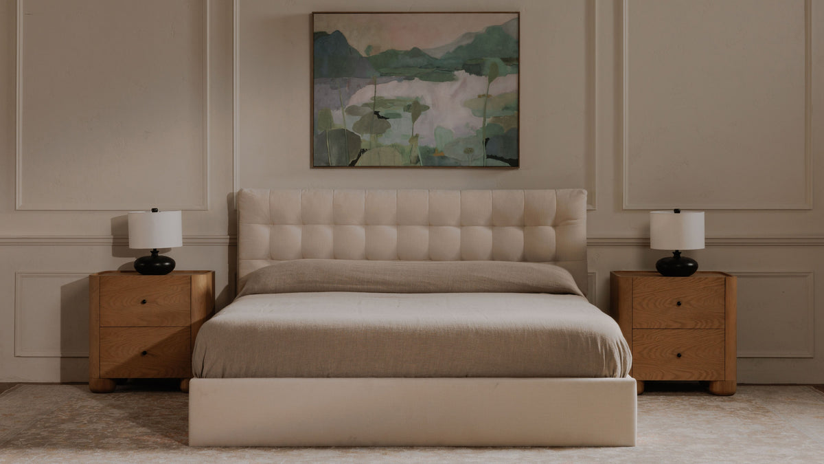 A pair of Moe's Home Collection Gibson nightstands in natural oak flanking a cream tufted bed in a panelled room, each carrying a black ceramic lamp, with a green lakeside painting hung above the headboard