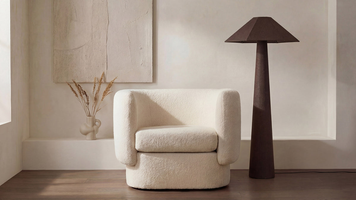 The Gigi floor lamp in dark brown linen and cotton, the same plaster-walled room as the cream frame, its tapered base and geometric shade reading as a dark silhouette beside a cream boucle swivel chair on an oak floor