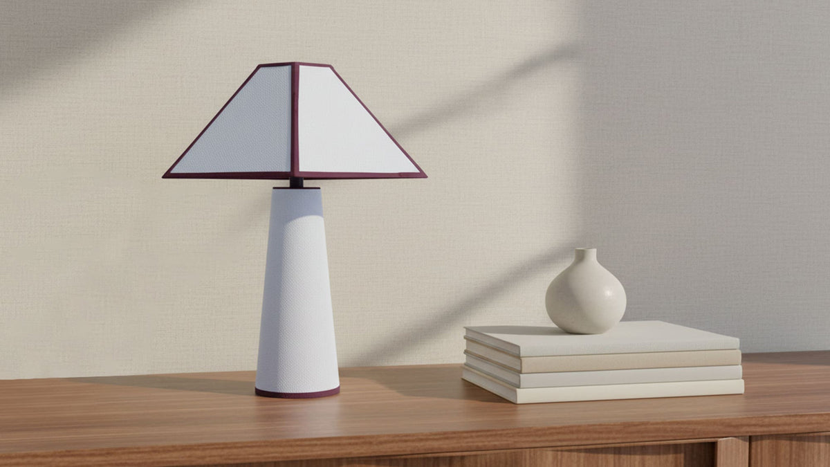 The Gigi table lamp in light blue, its fabric-wrapped conical base and wide geometric shade trimmed in contrast piping, on a walnut console beside a small white vase resting on stacked books