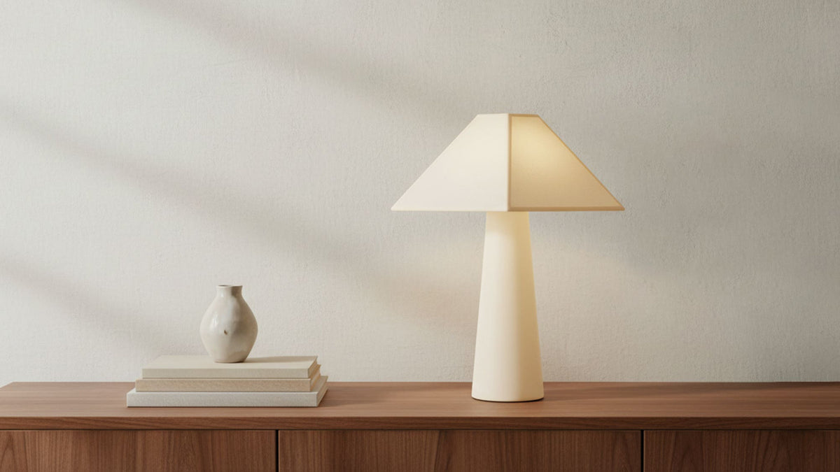 The Gigi table lamp in cream on a walnut console, its cotton-wrapped base and geometric shade catching low sunlight against a plaster wall, books and a small vase alongside