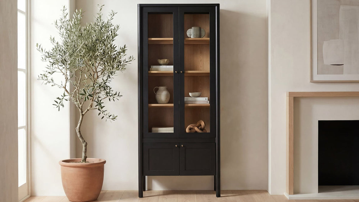 Moe's Home Collection Hannah tall cabinet in black, glass doors over a pale interior of ceramics, beside an olive tree and a stone fireplace
