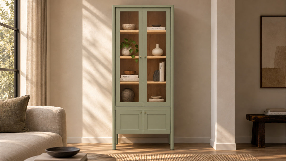 Moe's Home Collection Hannah tall cabinet in green, glass doors over an oak interior of bowls and books, in a sunlit room beside a pale sofa