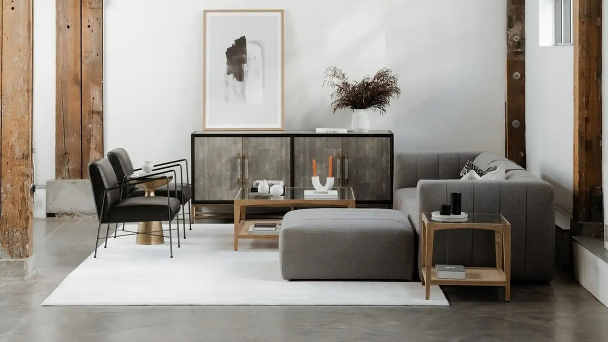 Moe's Home Collection Harrington square coffee table in solid oak with a tempered glass top and woven cane shelf, holding books in a white loft living room with exposed timber posts and a grey sectional