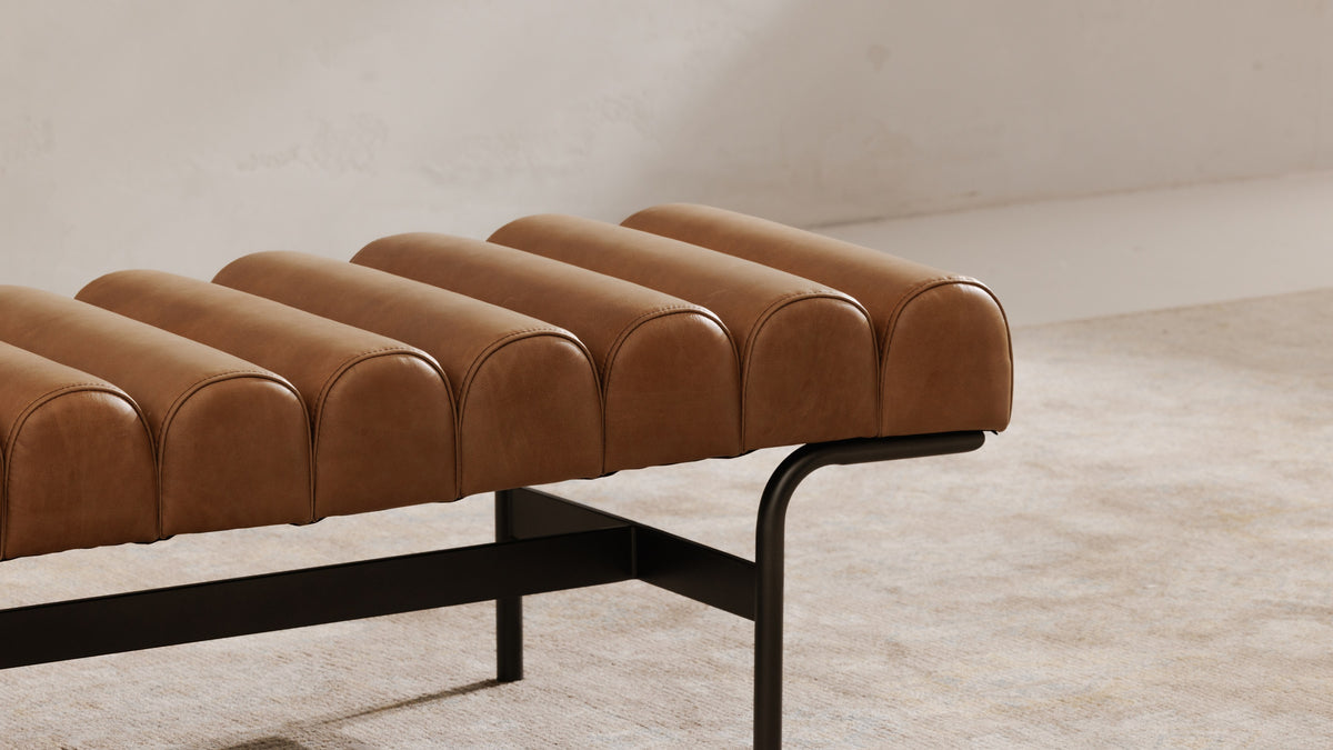 Close view of the Harrison bench in tan leather, showing the rounded leather channels running across the seat and the slim black iron frame beneath