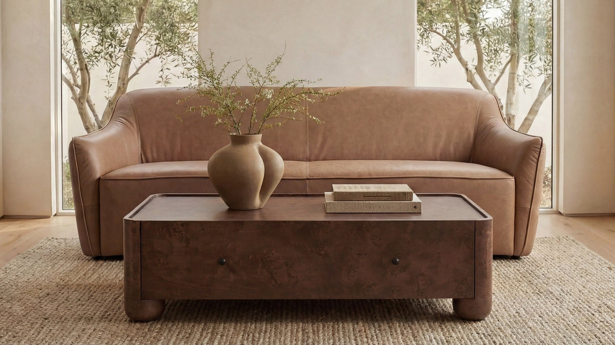 Moe's Home Collection Hawkins sofa in tan top-grain leather, its sloped arms and tall back seen straight on in a plaster-walled room between tall windows, behind a low burl-wood coffee table on a jute rug