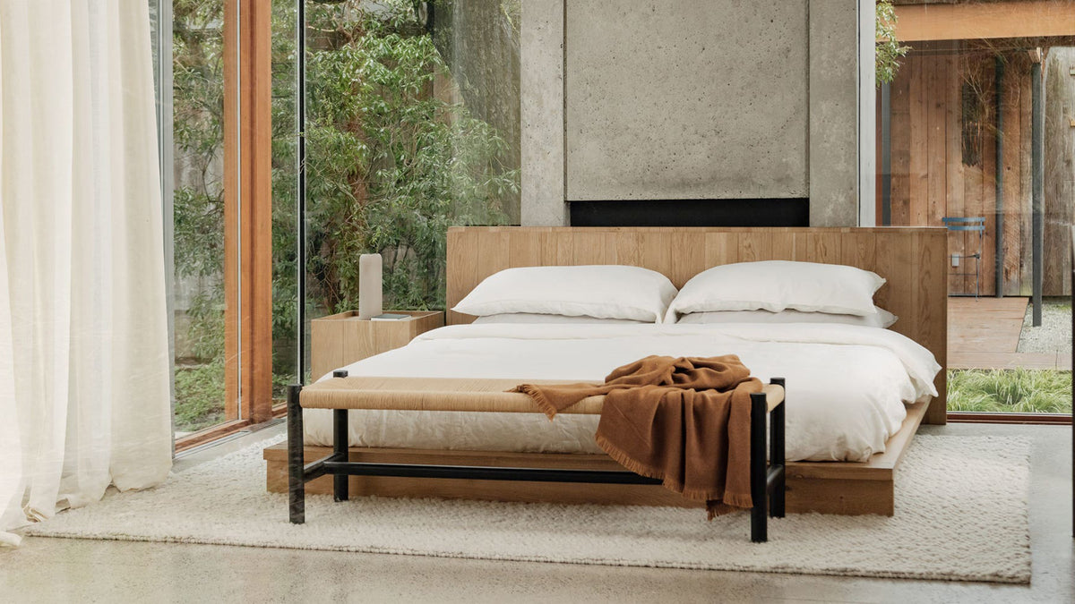 Moe's Home Collection Hawthorn bench with a black elm frame and woven natural-fibre rope seat, at the foot of a bed in a glass-walled bedroom, a rust throw folded across one end