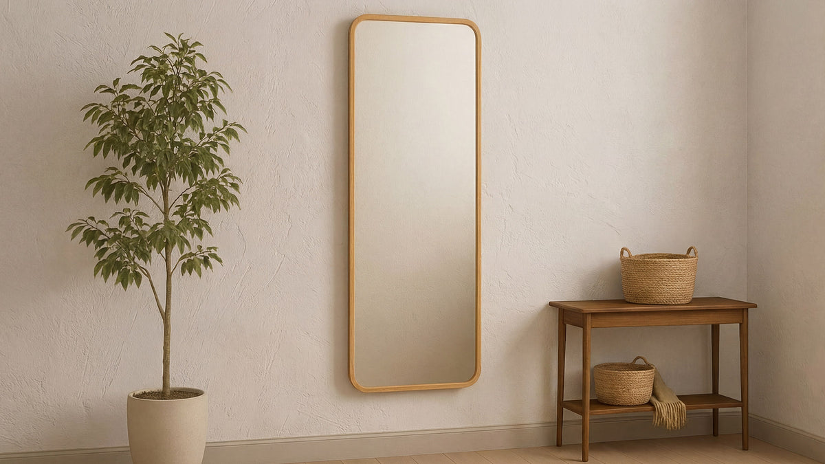 The Kai floor mirror in oak against a pale wall, a potted tree to one side and a wooden bench with baskets to the other