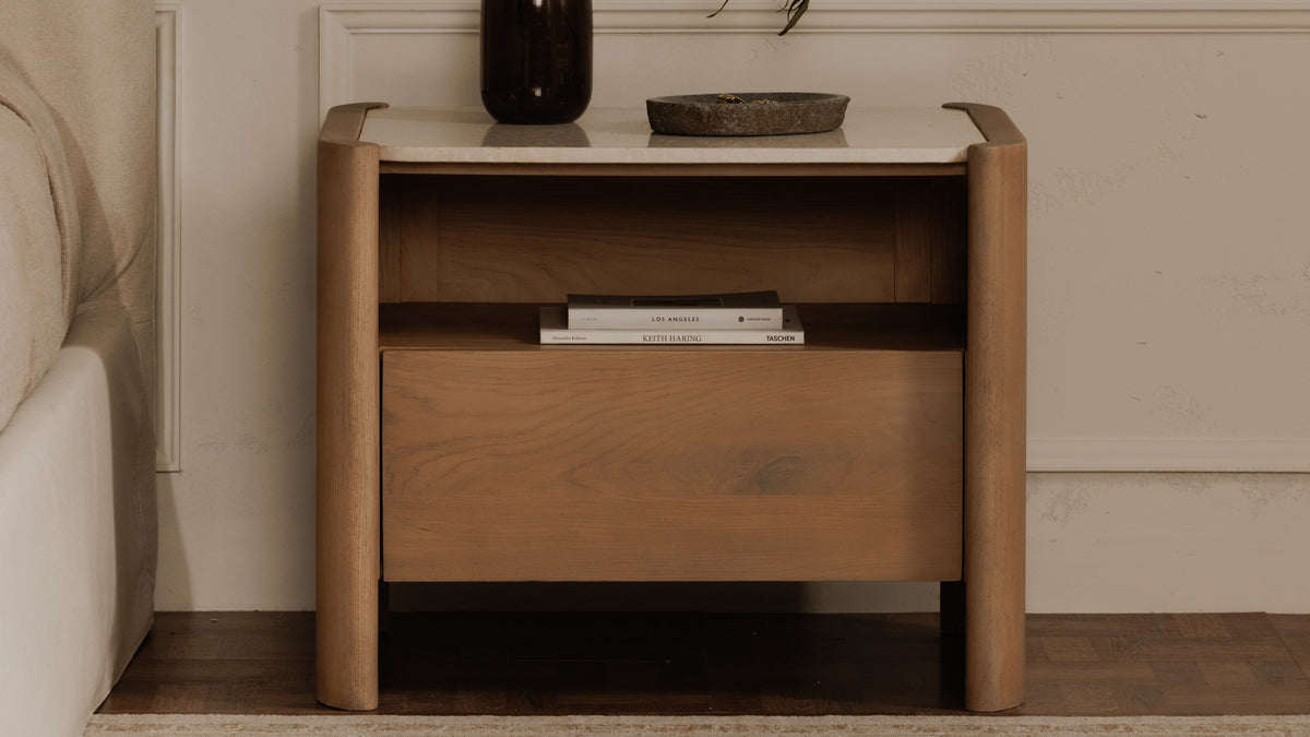 Moe's Home Collection Lane nightstand in natural oak beside an upholstered bed, its inset Bottichino marble top carrying a dark glass vase and a stone dish, with books resting in the open cubby above the closed drawer