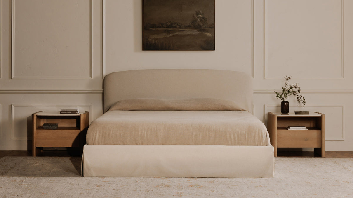 A pair of Moe's Home Collection Lane nightstands in natural oak flanking a curved linen bed in a panelled room, each with a pale marble top and an open cubby above one drawer, beneath a dark landscape painting