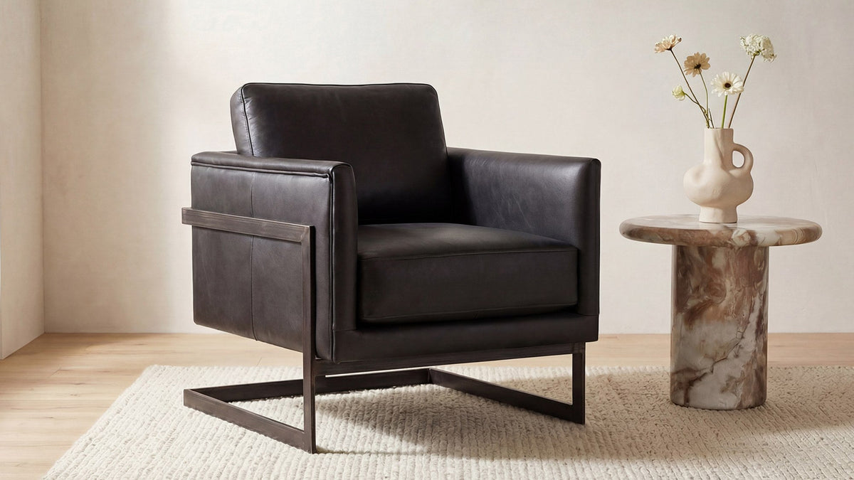 The Moe's Home Collection Luxley club chair in black aniline-dyed top-grain leather on a semi-cantilevered gunmetal iron base, on a woven wool rug beside a marble drum side table holding a pale stoneware vase of cream and ochre flowers
