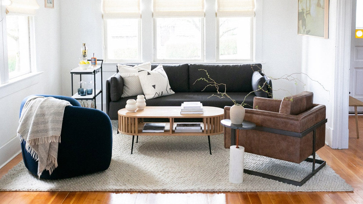 The Luxley club chair in brown leather on a black iron base, in a white-walled living room with a charcoal sofa, a navy swivel chair, a slatted oak coffee table and a jute rug on oak floorboards
