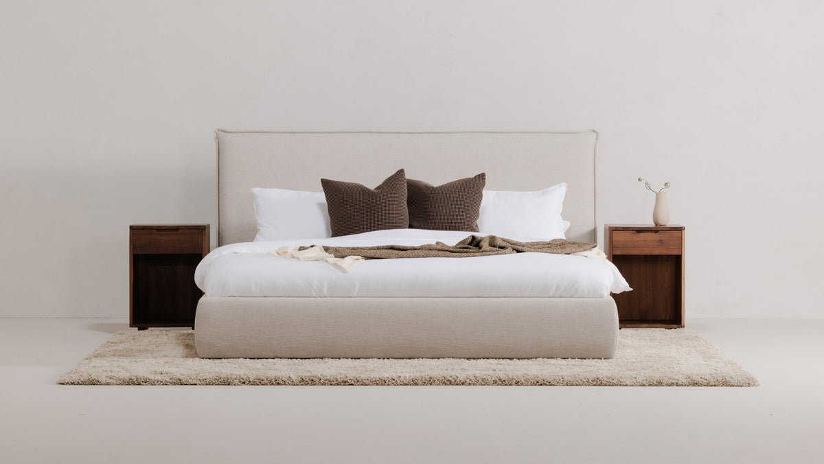 Moe's Home Collection Luzon bed in Wheat linen-blend upholstery, made up in white bedding with brown cushions on a cream shag rug, between two walnut nightstands
