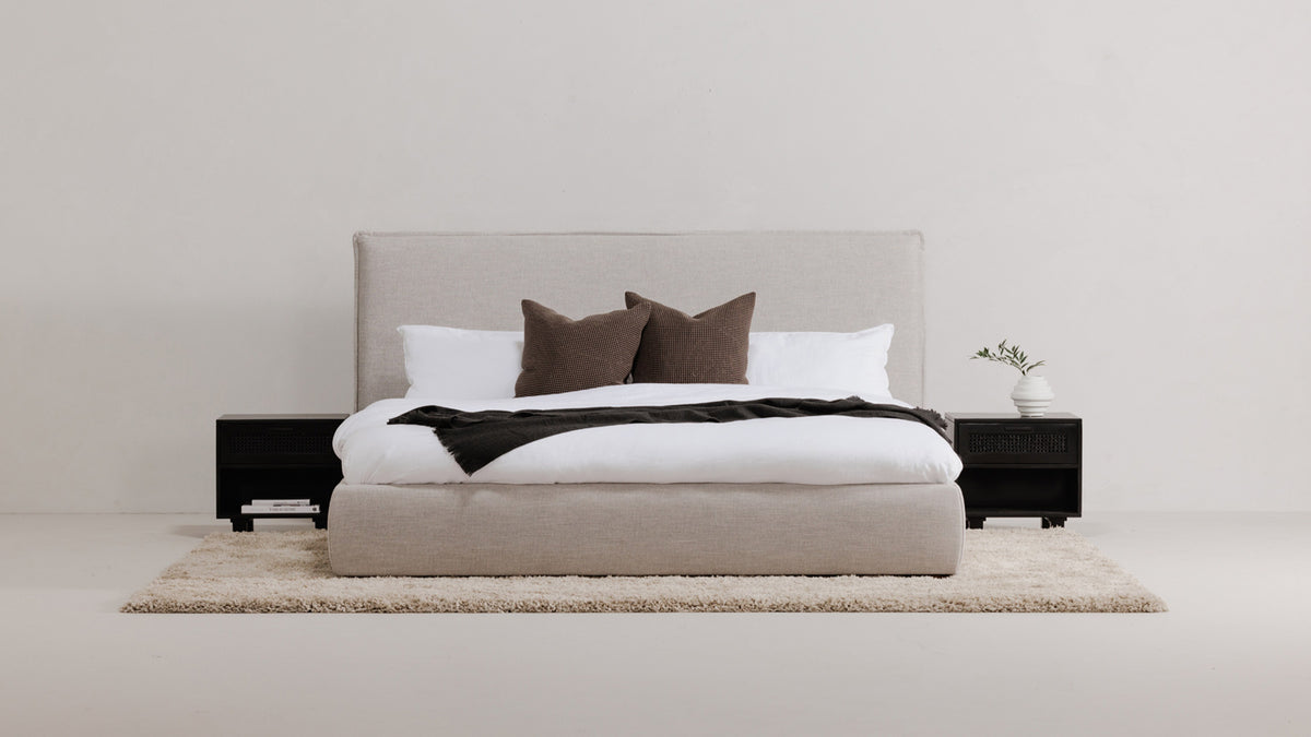 The Luzon bed in Light Gray linen-blend upholstery, made up in white bedding with a charcoal throw, between two black nightstands on a pale rug