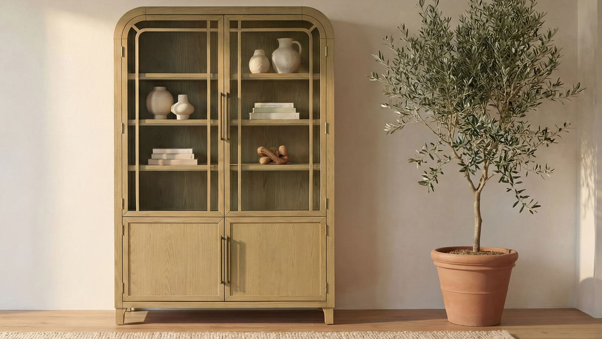 Moe's Home Collection Maddie cabinet in natural oak, tall with glass doors, holding ceramics and books, beside a potted olive tree