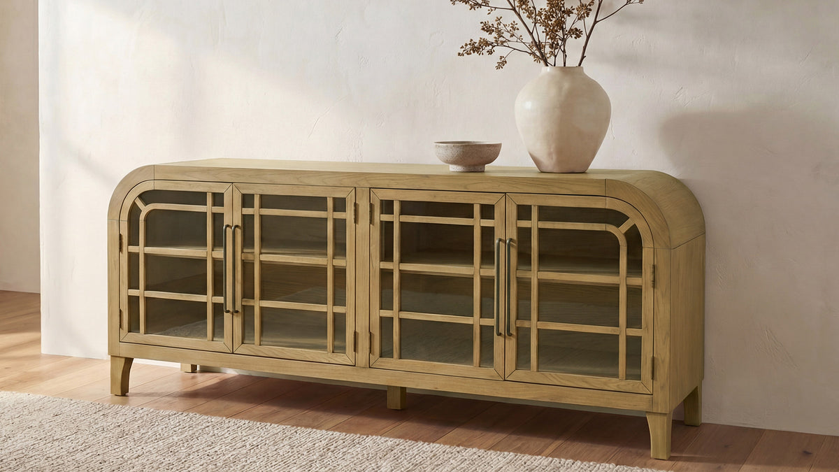 Moe's Home Collection Maddie sideboard in natural oak on a pale rug, its arched glass doors and fine mullions showing the shelves inside, with a tall vase of dried stems and a small bowl on top