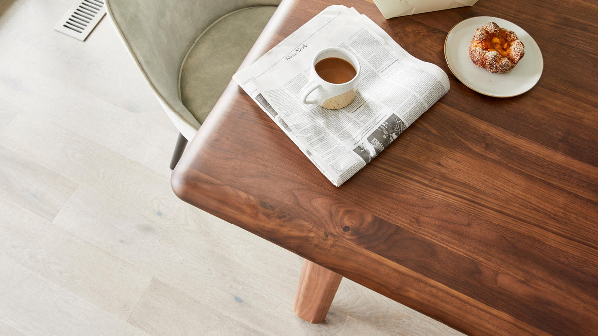 Overhead view of the Malibu dining table in solid walnut, its bullnose corner and one round leg in frame, with a coffee cup on a folded newspaper and a pastry on a plate