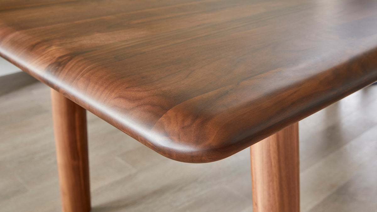 Close view of the Malibu dining table in solid walnut, showing the bullnose edge running around the corner and one round tapered leg on a pale wood floor