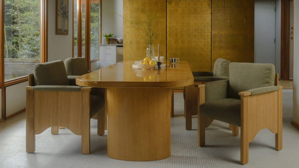 The Mallory dining table seen end on, its domed end and curved panel base surrounded by upholstered armchairs in a warm room with a gold tiled wall and a mosaic tile floor